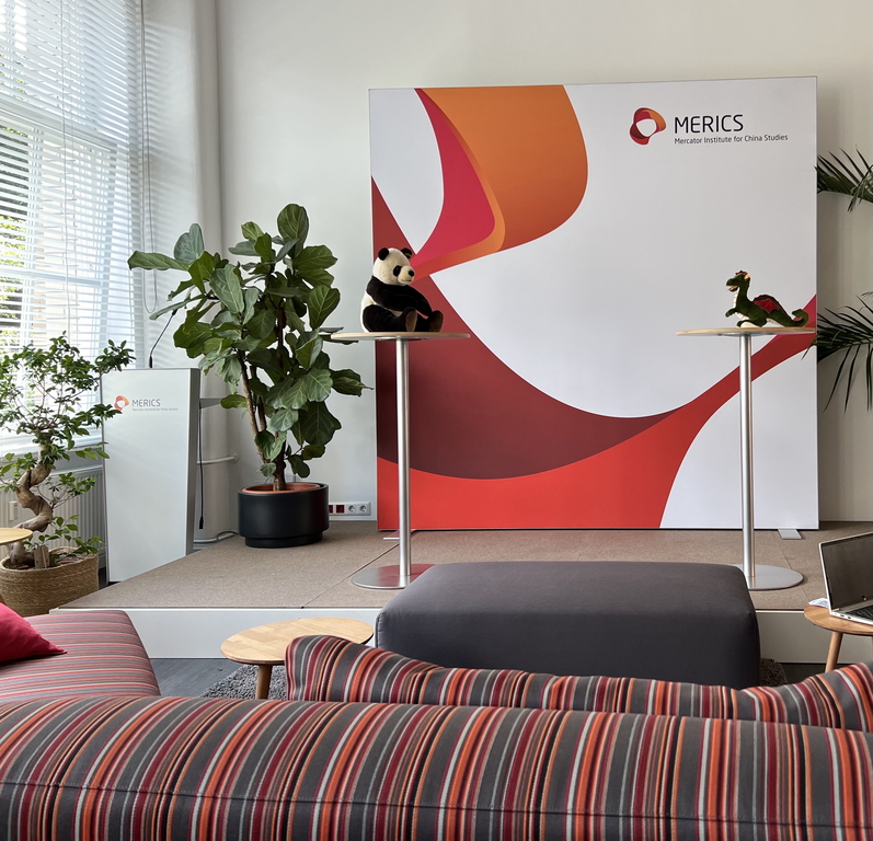 Ścianka z logotypami MERICS, przed którą stoją dwa wysokie stoliki koktajlowe. Na jednym stoliku leży maskotka pandy, a na drugim maskotka smoka.