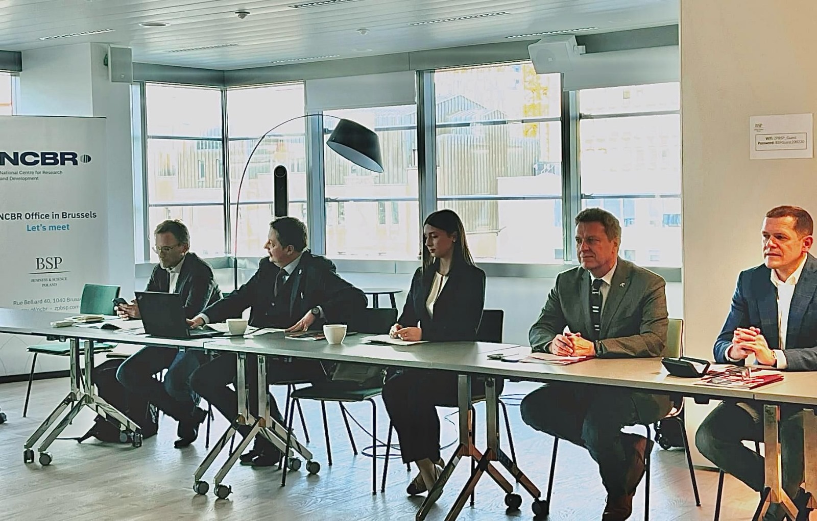 Pięć osób (czterech mężczyzn i jedna kobieta) siedzą przy długim stole na tle okien