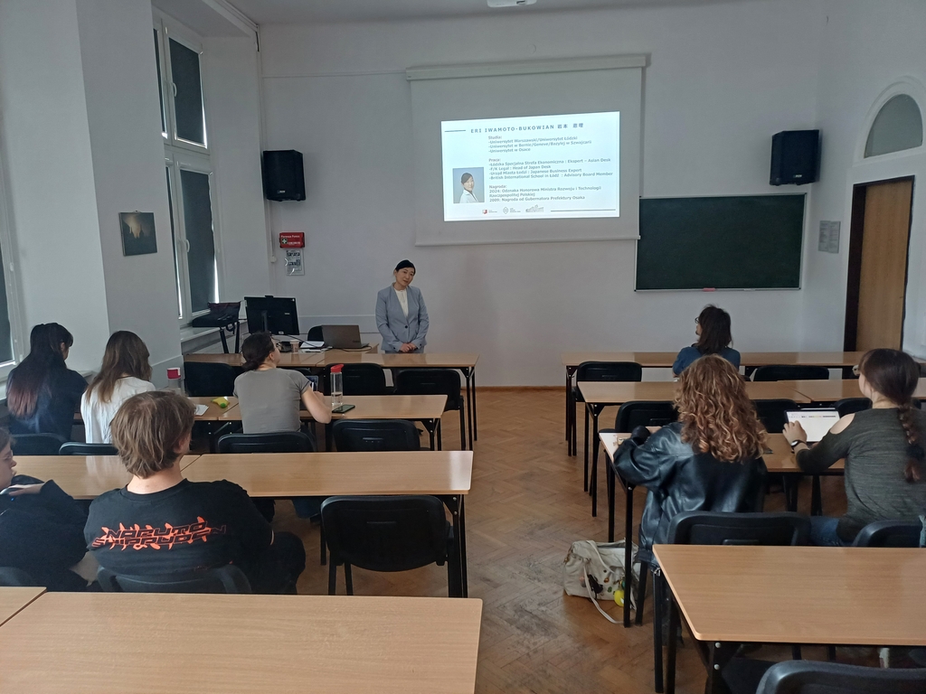 Prelegentka stojąca przed salą wykładową, prezentacja wyświetlana na ekranie z informacjami o jej doświadczeniu zawodowym; kilku studentów siedzi w ławkach i słucha.