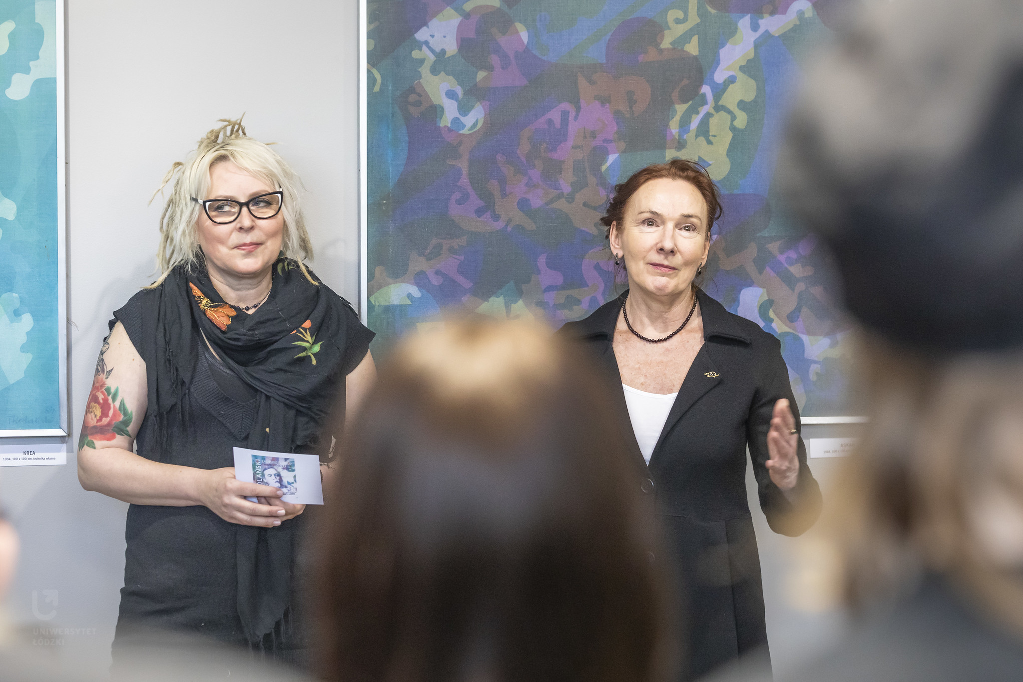 Na zdjęciu widoczne są dwie kobiety. Stoją twarzami do nas. Za nimi widczone są wiszące na ścianie obrazy. Kobiety widzimy od pasa w górę. 