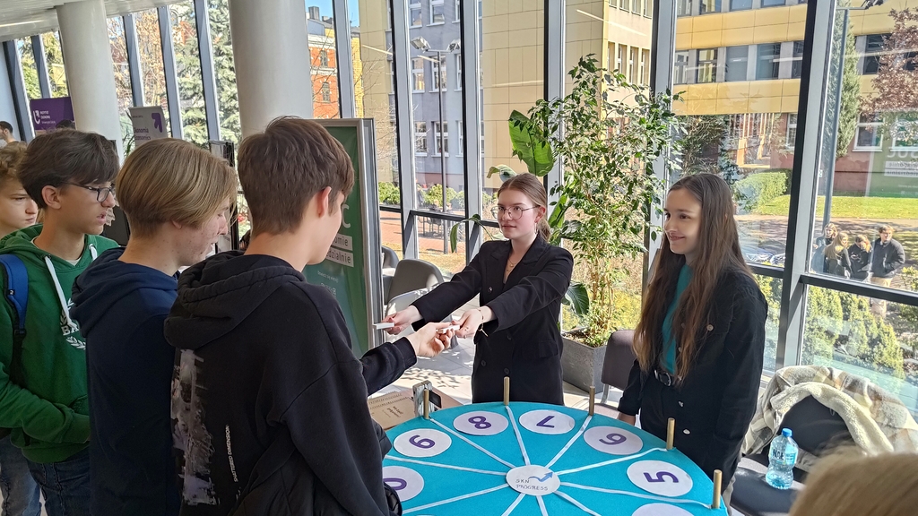 drzwi otwarte 2025 - dwie studentki SKN Progress rozdają nagrody uczniom ustawionym w kolejce przed stoiskiem EKO EkSoc