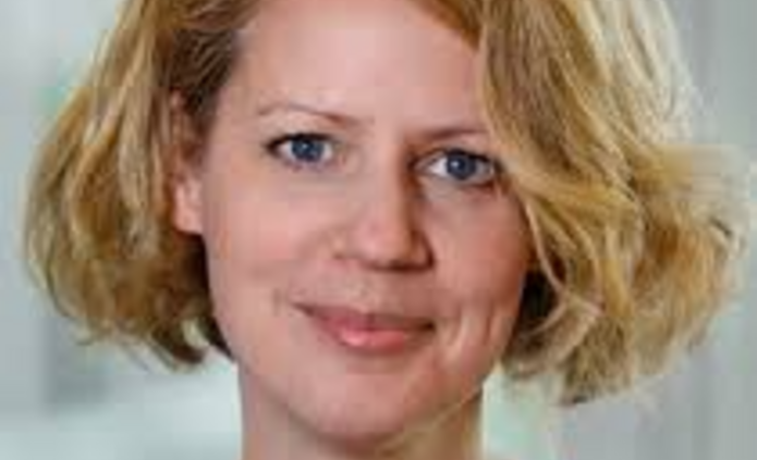 Portrait of a woman with short, wavy blonde hair wearing a light blue blouse, standing indoors against a softly blurred background and smiling gently at the camera.