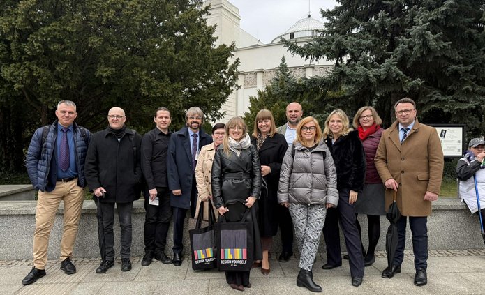 grupa ludzi stojąca przed budynkiem sejmu RP, są to kobiety i mężczyzni, stoją na chodniku, w tle widoczny budynek sali plenarnej sejmu.