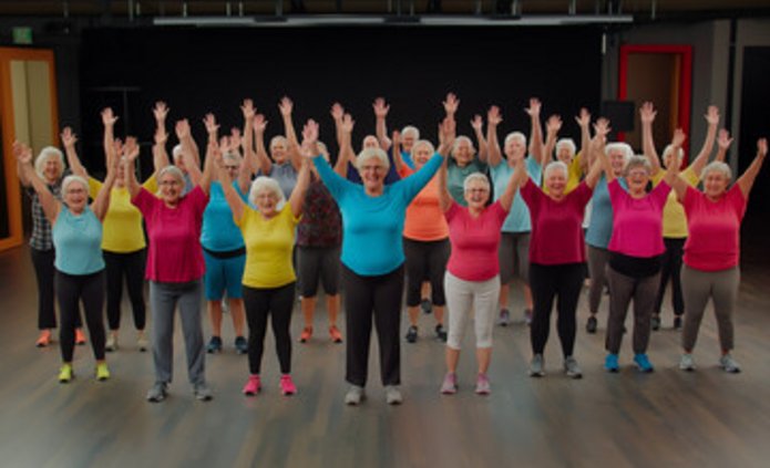 Grupa seniorów stoi na scenie. Są w kolorowych strojach sportowych. Mają podniesione ręce. Uśmiechają się.