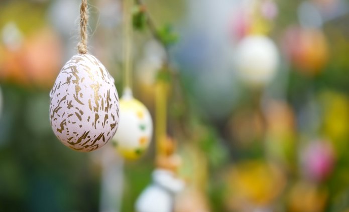 Zbliżenie na wielkanocną dekorację: białą pisankę z nieregularnym złotym wzorem, zawieszoną na sznurku. Jajko znajduje się po lewej stronie obrazu i jest ostre oraz dobrze widoczne. W tle, które jest rozmyte, wiszą inne kolorowe pisanki zawieszone na gałązkach. Widać pastelowe kolory – żółty, różowy, zielony i biały. Całość tworzy wiosenną, świąteczną atmosferę z dominującymi zielonymi i żółtymi odcieniami tła.