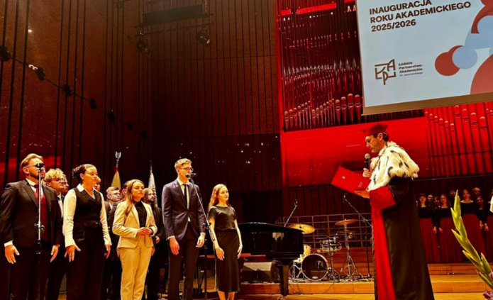 W dużej sali z podestem i organami w tle odbywa się uroczystość inauguracji roku akademickiego. Po prawej stronie stoi dostojna postać w todze i futrzanej pelerynie, trzymająca mikrofon i czytająca przemówienie z czerwonej teczki. Po lewej, na scenie przed mikrofonami, ustawiona grupa młodych osób — studenci i studentki — w eleganckich strojach, stoją prosto i poważnie. W tle widać część chóru oraz instrumentalistów, a nad sceną – baner z napisem „Inauguracja Roku Akademickiego 2025/2026”. Sceneria jest formalna, dekorowana czerwoną poświatą i przemyślanym oświetleniem, nadając wydarzeniu podniosły charakter.