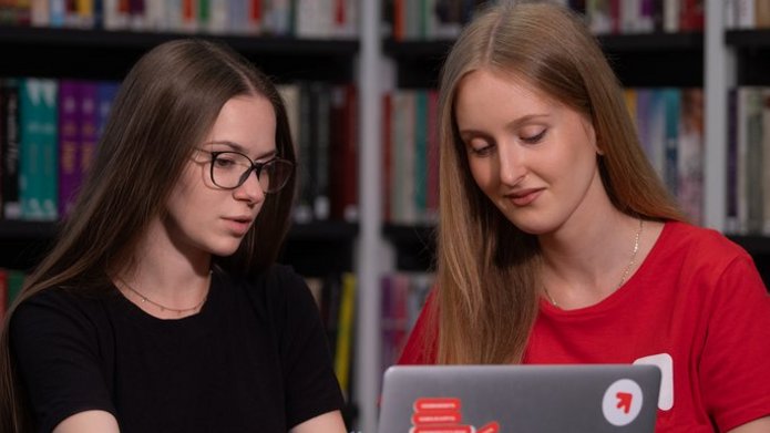 Dwie studentki siedzą przy laptopie w bibliotece i rozmawiają. 