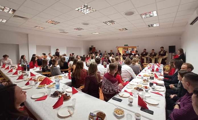 zdjęcie przedstawia zastawione stoły i osoby siedzące przy nich, zwrócone w stronę drzwi, gdzie grupa w strojach szlacheckich wykonuje kolędy