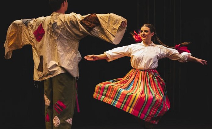 para tancerzy, partner stojący z rozłożonymi rękami na wzór stracha na wróble, przed nim tańczy partnerka w białej koszuli i pasiastej spódnicy