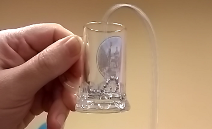 Na zdjęciu widać dłoń trzymającą niewielkie, przezroczyste szklane naczynie przypominające mały kieliszek. Wewnątrz naczynia znajdują się drobne, metaliczne kulki. Z prawej strony do naczynia wprowadzona jest przezroczysta, cienka rurka, która wygina się łukiem nad jego krawędzią i prowadzi na zewnątrz. Tło zdjęcia jest jednolite i jasne, w kolorze beżowym, dzięki czemu główny element – szklane naczynie z kulkami i rurką – jest dobrze widoczny na pierwszym planie. Całość wygląda jak fragment prostego doświadczenia fizycznego z użyciem małego naczynia, kulek i elastycznej rurki.