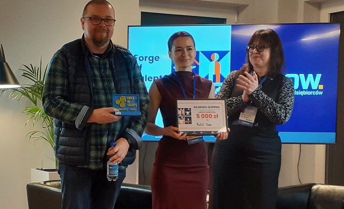 Na zdjęciu troje uczestników konkursu Forge of Talents 2025 stoi na czerwonym dywanie w sali konferencyjnej. Dwoje z nich trzyma dyplom i tabliczkę z napisem „Nagroda Główna 5000 zł”, a obok nich kobieta klaszcze, gratulując zwycięzcom. W tle widać ekran z logo wydarzenia Forge of Talents oraz sprzęt oświetleniowy.