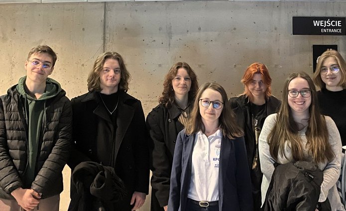 Na zdjęciu widać grupę dziewięciu osób stojących w dwóch rzędach na tle jasnej, betonowej ściany we wnętrzu budynku. Wszyscy patrzą w stronę aparatu i uśmiechają się. Osoby mają na sobie jesienne ubrania – kurtki, swetry i bluzy. W tle, po prawej stronie, znajduje się wejście oznaczone napisem „Wejście / Entrance” oraz kilka piktogramów z informacjami dla odwiedzających. Zdjęcie ma charakter pamiątkowy i przedstawia uczestników wydarzenia stojących wspólnie w grupie.