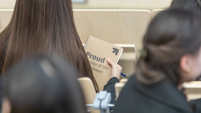 Na zdjęciu widać grupę osób siedzących tyłem do nas. Jedna z nich trzyma w dłoniach notes w kolorze beżowym, na którym jest napis proud member of the unilodz team