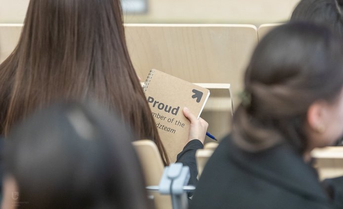 Na zdjęciu widać grupę osób siedzących tyłem do nas. Jedna z nich trzyma w dłoniach notes w kolorze beżowym, na którym jest napis proud member of the unilodz team