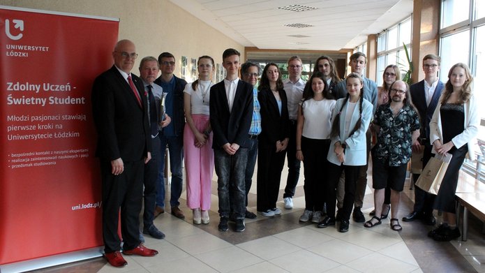 Grupa młodych i starszych osób stojących w korytarzu budynku. Wszyscy są elegancko ubrani, niektórzy z nich trzymają dokumenty. Po lewej stronie widoczny jest czerwony roll-up z napisem "Zdolny Uczeń - Świetny Student" . W tle widać okna, przez które wpada naturalne światło.