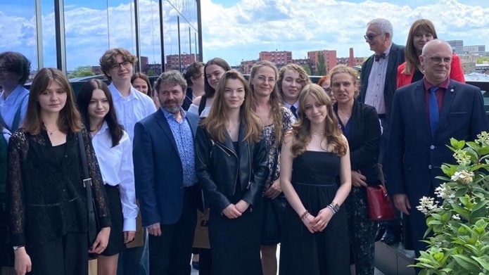 Grupa osób stojących na zewnątrz na tarasie lub balkonie budynku. Wszyscy są elegancko ubrani. Wśród grupy znajduje się zarówno młodzież, jak i dorośli. Niektóre osoby trzymają torby z logo Uniwersytetu Łódzkiego. W tle widać fragmenty miejskiego krajobrazu oraz zieleń.