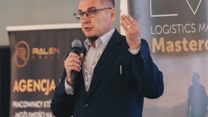 mężczyzna przemawiający przed publicznością, a za nim standy