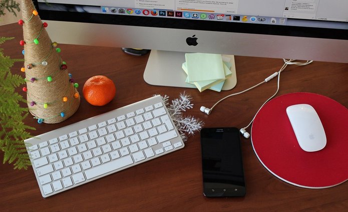 Na zdjęciu widoczne jest biurko z drewnianym blatem. Na środku stoi monitor komputerowy marki Apple. Przed monitorem znajduje się biała klawiatura oraz biała mysz komputerowa na czerwonej podkładce. Po lewej stronie biurka ustawiona jest dekoracyjna choinka wykonana ze sznurka, ozdobiona kolorowymi światełkami. Obok niej leży mandarynka. Na blacie widoczny jest także czarny smartfon, białe słuchawki przewodowe oraz kilka karteczek samoprzylepnych. Całość tworzy świąteczną, biurową aranżację miejsca pracy.