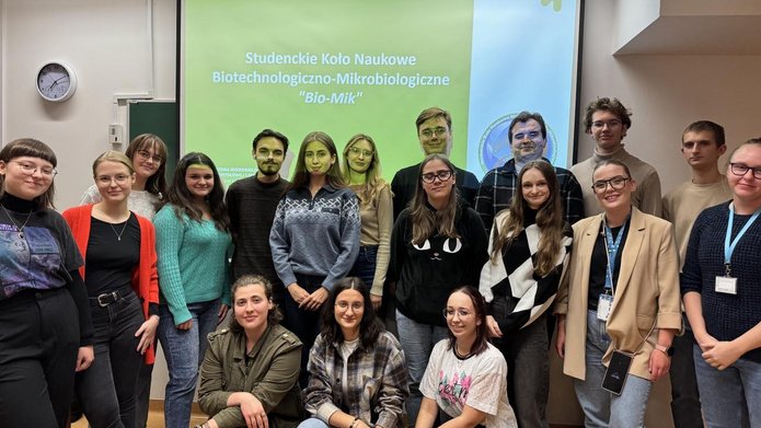 Zdjęcie uczestników zebrania inaugarcyjnego SKN Biotechnologiczno-Mikrobiologicznego. Przedstawia 18 osób na tle slajdu zatytułowanego "Studenckie Koło Naukowe Biotechnologiczno-Mikrobiologiczne "Bio-Mik"".