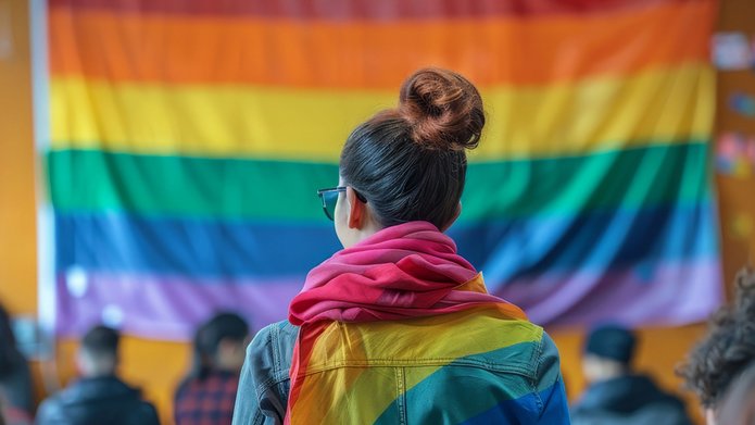 Osoba z tęczową flagą na ramionach widziana od tyłu na tle tęczowej flagi