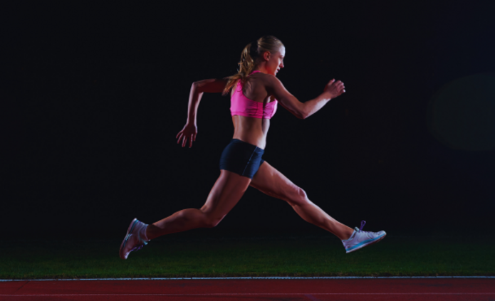 Na ciemnym tle widać młodą sportsmenkę w różowym topie i czarnych spodenkach, która dynamicznie biegnie po bieżni lekkoatletycznej. Jej sylwetka uchwycona jest w locie, co podkreśla siłę i energię ruchu.