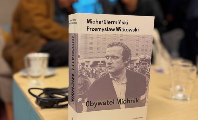 Zdjęcie ze spotkania, okładka nas stole, w tle Adam Michnik