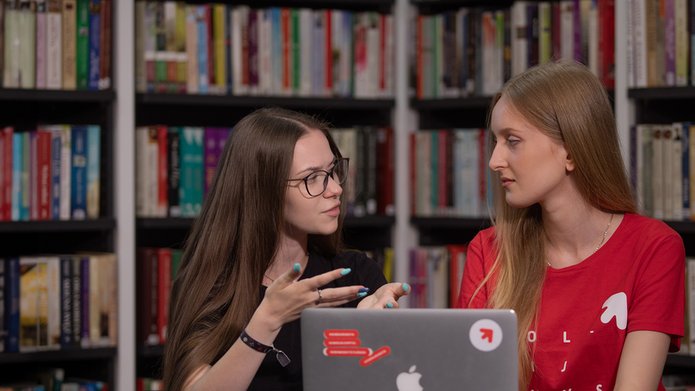 kolorowe zdjęcie przedstawiające dwie studentki, które na tle regałów z książkami wspólnie konsultują realizację projektu