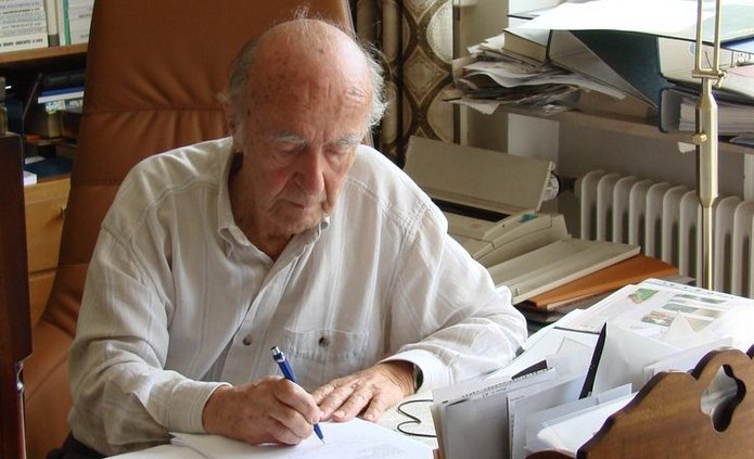 Karl Dedecius siedzi przy biurku z dużą liczbą papierów, książek i notatek, pisząc długopisem na kartce. Za nim znajduje się wysoki regał wypełniony książkami, a z boku wpada światło dzienne z okna. Scena przedstawia spokojną, skupioną pracę w otoczeniu pełnym materiałów naukowych.
