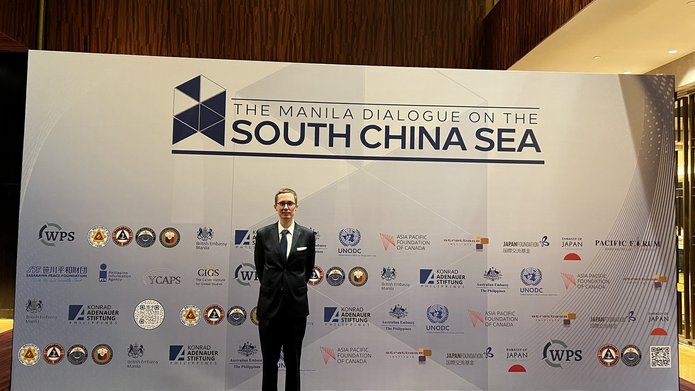 Mężczyzna w garniturze stoi przed ścianą z logotypami i napisem „The Manila Dialogue on the South China Sea” podczas konferencji w Manili.