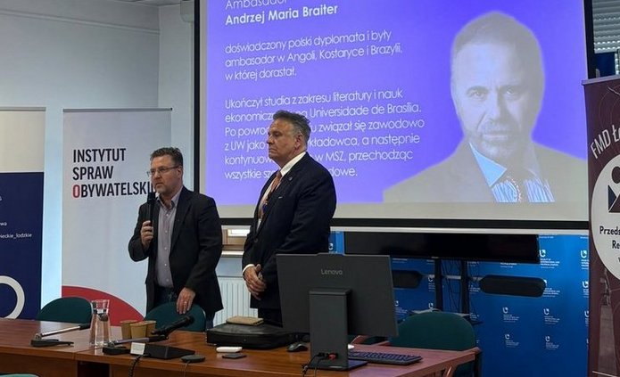 Osoba stojąca przy stole konferencyjnym przemawia do mikrofonu w sali spotkań, obok innej osoby stojącej przodem do zgromadzonych. W tle widoczny jest ekran z prezentacją zawierającą tekst: „Ambasador Andrzej Maria Braiter” oraz opis dotyczący polskiego dyplomaty i logo „Ministerstwo Spraw Zagranicznych”. Po lewej stronie ustawione są roll-upy, w tym „Instytut Spraw Obywatelskich”, a po prawej baner „FMD Łódź – Przedstawicielstwo Regionalne w Łodzi”. W pomieszczeniu znajdują się stoły konferencyjne, zielone krzesła, komputer Lenovo, mikrofony oraz naczynia z napojami. Nad salą widać sufit podwieszany z oświetleniem i zamontowany projektor.