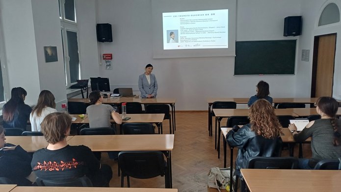Prelegentka stojąca przed salą wykładową, prezentacja wyświetlana na ekranie z informacjami o jej doświadczeniu zawodowym; kilku studentów siedzi w ławkach i słucha.