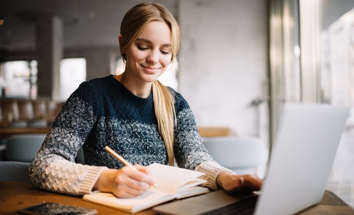 Young attractive university student using laptop computer, studying at modern library. Cheerful caucasian woman writes notes, planning working process, sitting in loft cafe. Exam preparation. Studentka pracuje przy komputerze w czytelni