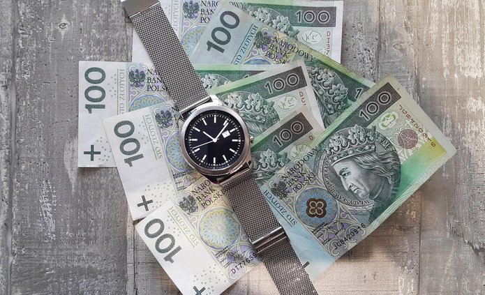 Na zdjęciu widoczny jest zegarek naręczny z metalową, srebrną bransoletą i czarną tarczą, położony na rozłożonych banknotach o nominale 100 złotych. Banknoty są ułożone wachlarzowo i częściowo nachodzą na siebie. Na banknotach widoczny jest wizerunek historycznej postaci oraz zielono-niebieska kolorystyka charakterystyczna dla polskich stu złotych. Tło stanowi jasna, drewniana powierzchnia o widocznej fakturze. Zdjęcie symbolicznie łączy temat pieniędzy i czasu.