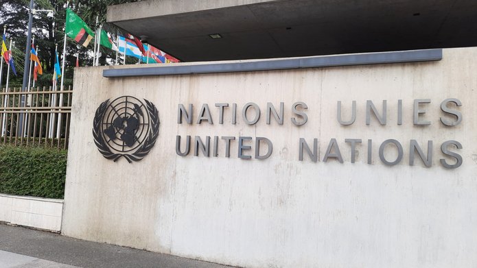 Zbliżenie na betonową ścianę z logo i napisem „Nations Unies – United Nations” przy wejściu do siedziby ONZ w Genewie, z widocznymi flagami państw członkowskich.