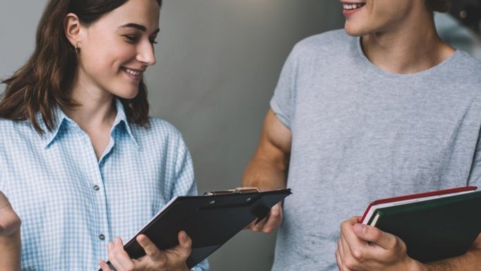 Dwoje uśmiechniętych studentów stoi obok siebie — jeden trzyma notesy, drugi podkładkę na dokumenty.