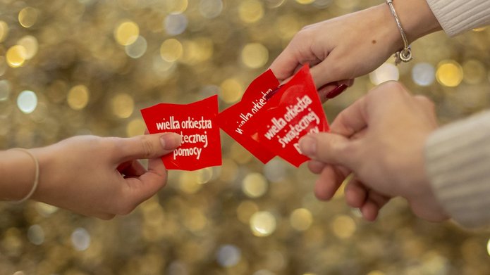3 ręce trzymające czerwone serduszka z napisem Wielka Orkiestra Świątecznej Pomocy" na tle złotych monet