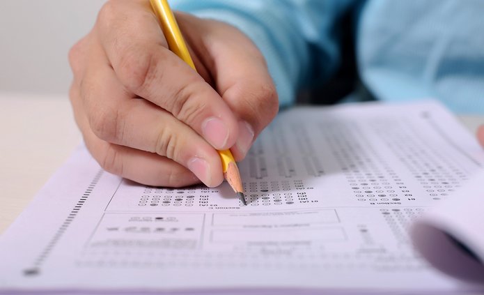 na kartce papieru są zadania egzaminacyjne i widać męską dłoń z długopisem rozwiązującą zadania