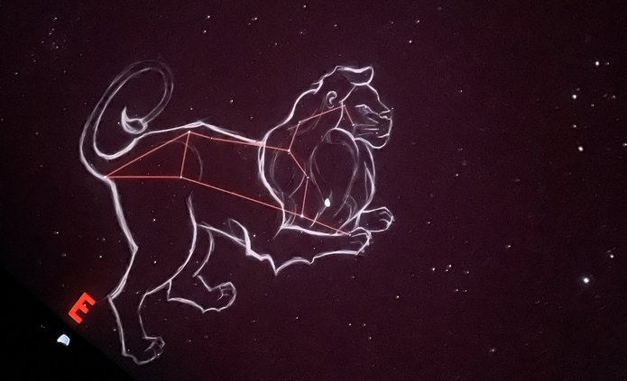 Na ciemnym, gwiaździstym tle widoczna jest ilustracja konstelacji Lwa. Gwiazdy połączone są cienkimi, czerwonymi liniami tworzącymi charakterystyczny kształt, a całość otacza jasny, szkicowy kontur przedstawiający lwa w pozycji stojącej. Po lewej stronie znajduje się czerwona litera „E”, oznaczająca kierunek wschodni. Tło jest jednolicie ciemne, a rozproszone punkty światła przedstawiają gwiazdy.