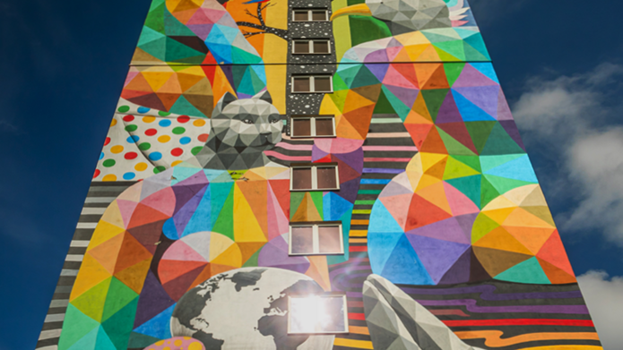 Na zdjęciu znajduje się wielobarwny mural pokrywający całą fasadę wysokiego budynku. Obraz składa się z geometrycznych, wielościennych form tworzących postacie ludzi i zwierząt — widoczne są m.in. delfin, ptaki, niedźwiedź i głowa człowieka w profilu. Elementy są pomalowane w żywe, kontrastowe kolory tworzące mozaikę w stylu low poly. Centralnie przez mural biegną okna w pionowym rzędzie, a w tle widoczne jest niebieskie niebo z lekkim zachmurzeniem. Mural przedstawia różnorodność biologiczną i jedność ludzi z naturą, tworząc symboliczny i optymistyczny przekaz.