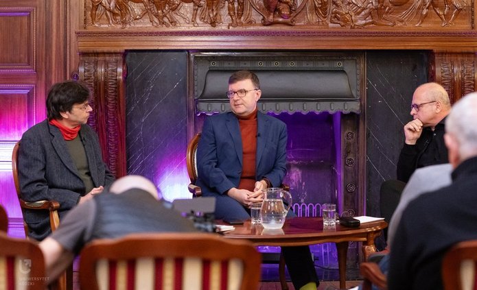 Na zdjęciu widać trzech mężczyzn. Mężczyźni siedzą przy stoliku, rozmawiają ze sobą. Są w sali, która wygląda jak sala balowa. Za nimi widać kominek, w którym nie pali się ogień. 