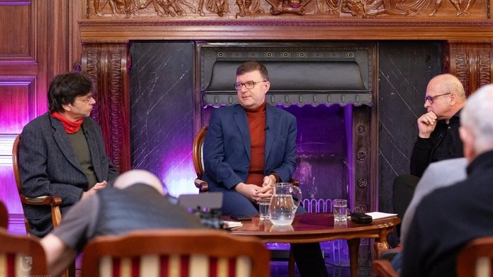 Na zdjęciu widać trzech mężczyzn. Mężczyźni siedzą przy stoliku, rozmawiają ze sobą. Są w sali, która wygląda jak sala balowa. Za nimi widać kominek, w którym nie pali się ogień. 