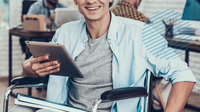 Uśmiechnięty mężczyzna na wózku inwalidzkim trzyma w dłoni tablet. W tle dwie inne osoby pracują przy komputerze.