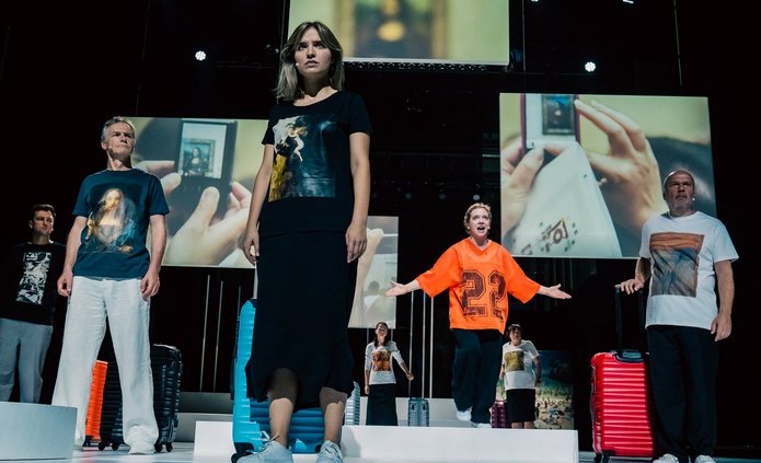 Na zdjęciu widać grupę osób stojących i patrzących w jeden wspólny punkt. To kobiety i mężczyźni ubrani jak turyści, koszulki sportowe spodnie i obuwie. Przy każdej osobie stoi walizka. W tle widoczne na wielkich ekranach zdjęcia z podróży, z turystami.