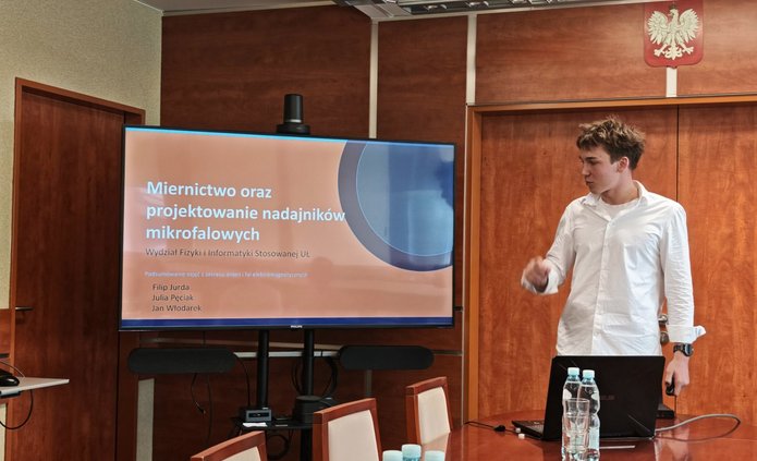 W sali konferencyjnej młody mężczyzna prowadzi prezentację przy dużym ekranie multimedialnym. Ubrany jest w białą koszulę i stoi po prawej stronie ekranu, zwrócony w jego kierunku. Na ekranie wyświetlony jest slajd tytułowy prezentacji zatytułowanej „Miernictwo oraz projektowanie nadajników mikrofalowych”. Przed prelegentem znajduje się stół konferencyjny z otwartym laptopem, butelkami wody, szklankami i talerzykiem z poczęstunkiem. W tle widoczne są drewniane ściany oraz godło Polski umieszczone wysoko nad drzwiami. Zdjęcie przedstawia formalne wystąpienie podczas wydarzenia edukacyjnego lub naukowego.