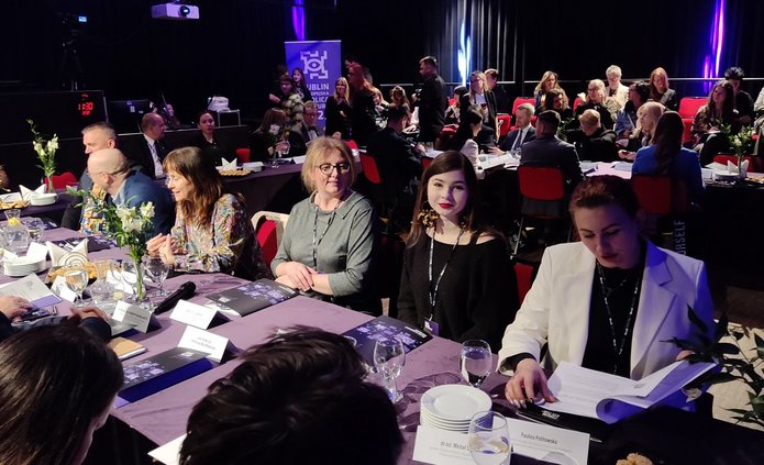 Na zdjęciu widoczne są osoby siedzące za stołem. Jest to spora grupa. Osoby debatują w sali, która wygląda jak sala teatralna. Jest dość ciemno, a oświetlenie jest teatralne, z fioletowym odcieniem. 