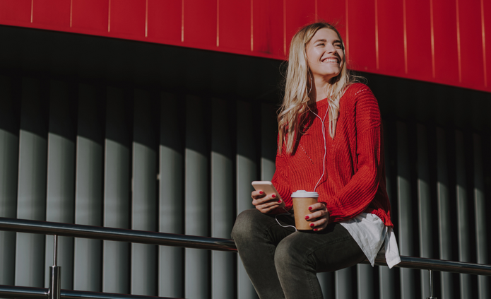 Na zdjęciu widoczna jest młoda kobieta siedząca na metalowej barierce. Ma na sobie czerwony sweter, ciemne spodnie i białą koszulę wystającą spod swetra. W jednej dłoni trzyma smartfon, w drugiej papierowy kubek z kawą; w uszach ma słuchawki. Kobieta uśmiecha się i patrzy w bok. Tło stanowi nowoczesna architektura z pionowymi, ciemnymi panelami oraz intensywnie czerwonym elementem konstrukcyjnym u góry kadru.