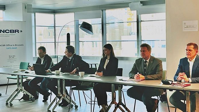 Pięć osób (czterech mężczyzn i jedna kobieta) siedzą przy długim stole na tle okien