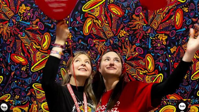 2 młode kobiety stojące na tle korowego tła z czerwonymi balonami w kształcie serca z napisem wielka orkiestra świątecznej pomocy