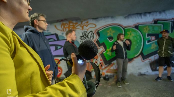 Na zdjęciu widać grupę ludzi. Stoją przejściu podziemnym. Trzymają w rękach urządzenia do nagrywania dźwięków. 
