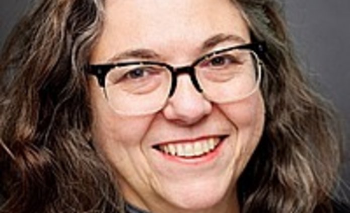 Studio headshot of a woman with long dark hair and glasses, wearing a dark blazer over a gray top, smiling in front of a neutral gray background.